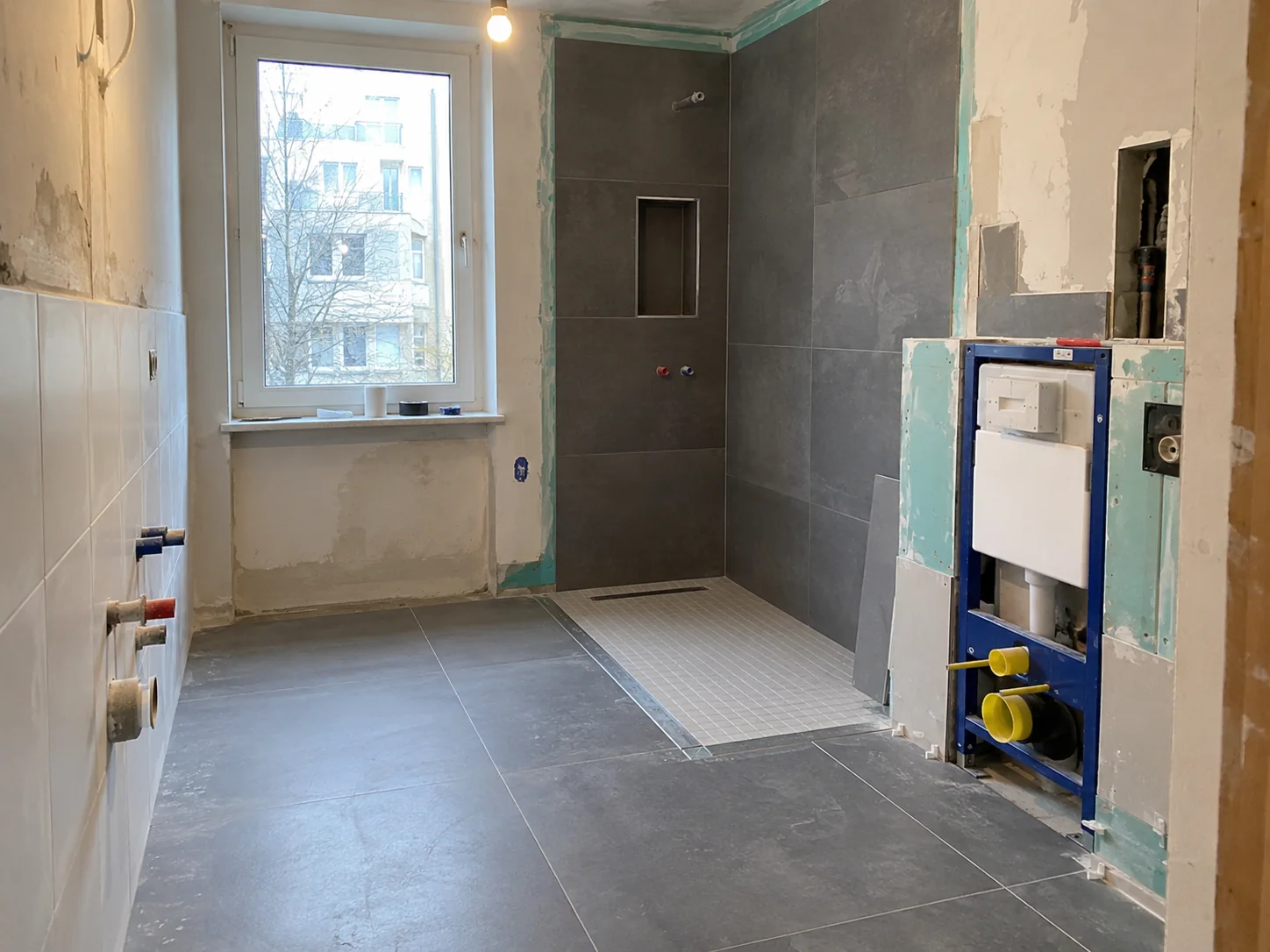 Badezimmer im Ausbau mit grauen Boden- und Wandfliesen, bodengleichem Duschbereich und noch unverkleidetem WC-Vorwandelement